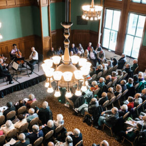 Aerial view of a classical music performance as part of the Courtroom Concerts series at Landmark Center in St. Paul. Musicians on stage include a violinist, cellist, and violist. An audience of seated people observes, surrounded by ornate wood paneling and large windows. A chandelier hangs overhead.