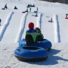 Two children wearing winter gear sit in blue snow tubes atop a snowy hill, ready to slide down multiple groomed lanes. Other people are seen tubing down the hill in the background, enjoying a snowy day out.