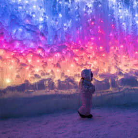 A child in winter clothing stands inside an ice cave, surrounded by vibrant pink, purple, and blue lights illuminating the icy walls, creating a magical atmosphere.
