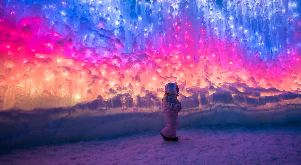A child in winter clothing stands inside an ice cave, surrounded by vibrant pink, purple, and blue lights illuminating the icy walls, creating a magical atmosphere.