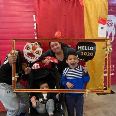 A group of people pose with a large picture frame in front of a red and yellow backdrop. One person wears a lion mask. Two kids hold signs; one says "Hello 2020." A baby is seated in a stroller. A banner reading "Cakes" hangs in the background.