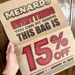 A hand holds a brown paper bag from Menards with bold red and black text advertising a 15% off bag sale promotion. The offer applies to everything that fits inside the bag, valid from January 1 to January 12, 2025.