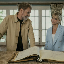 A man and a woman stand behind a large open book on a wooden desk. The man is wearing a beige jacket, and the woman is in a light blue blazer. They are indoors with large windows and patterned curtains in the background.