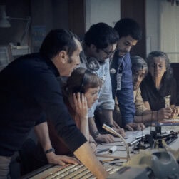 In a still scene from the movie SEPTEMBER 5, a group of six people stand around a desk in a dimly lit room, engaged in a serious discussion. Some wear headsets, and documents are scattered on the table. Photographs and equipment are visible in the background.