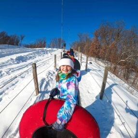 A young girl in a colorful jacket and winter hat enjoys snow tubing on a bright, sunny day. She is sitting on a red tube, being pulled up a snowy hill against a backdrop of trees and a clear blue sky.